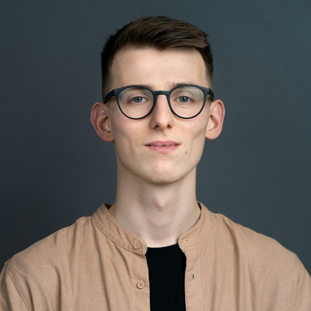 Portrait of a young man wearing glasses and a button down shirt against a gray background.