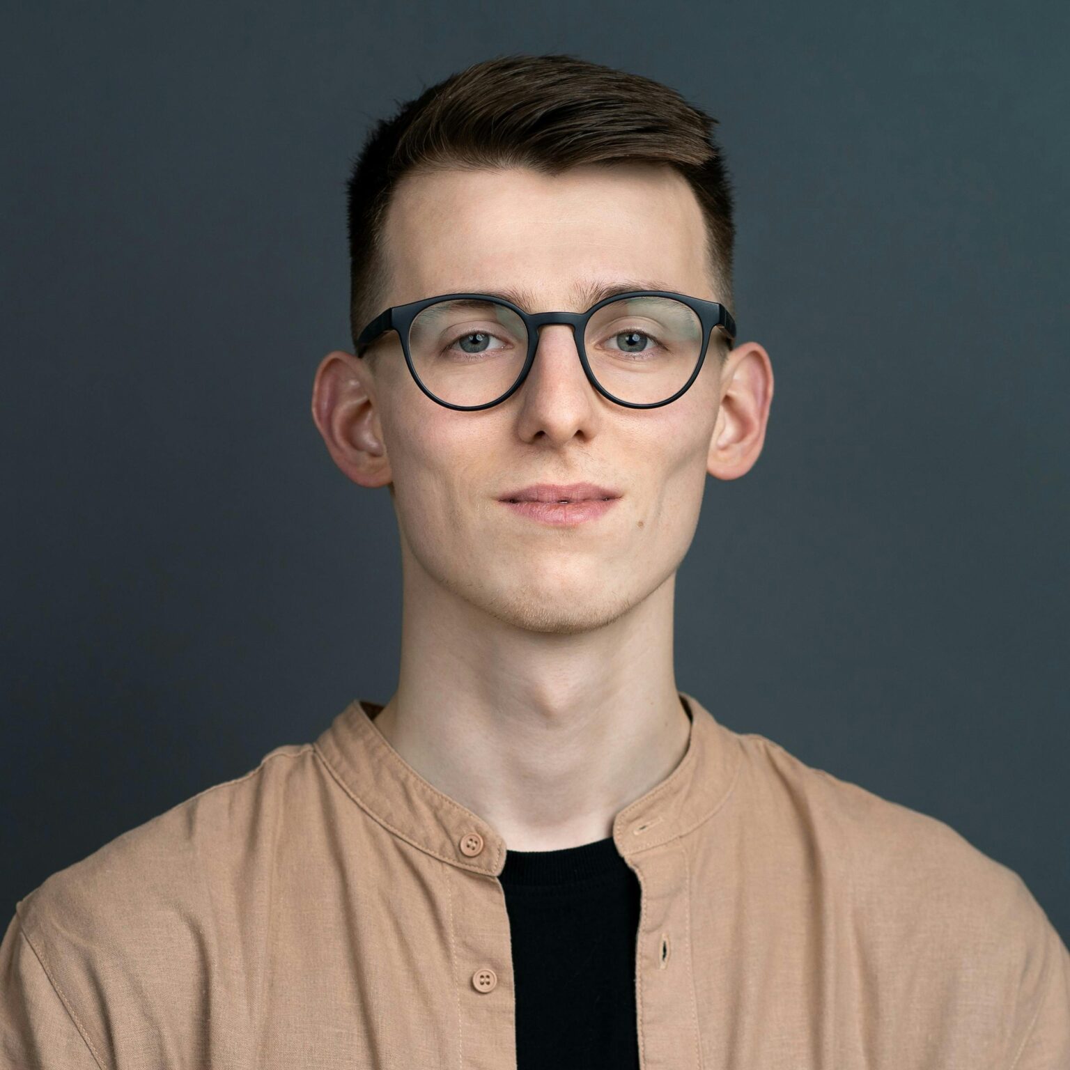 Portrait of a young man wearing glasses and a button down shirt against a gray background.